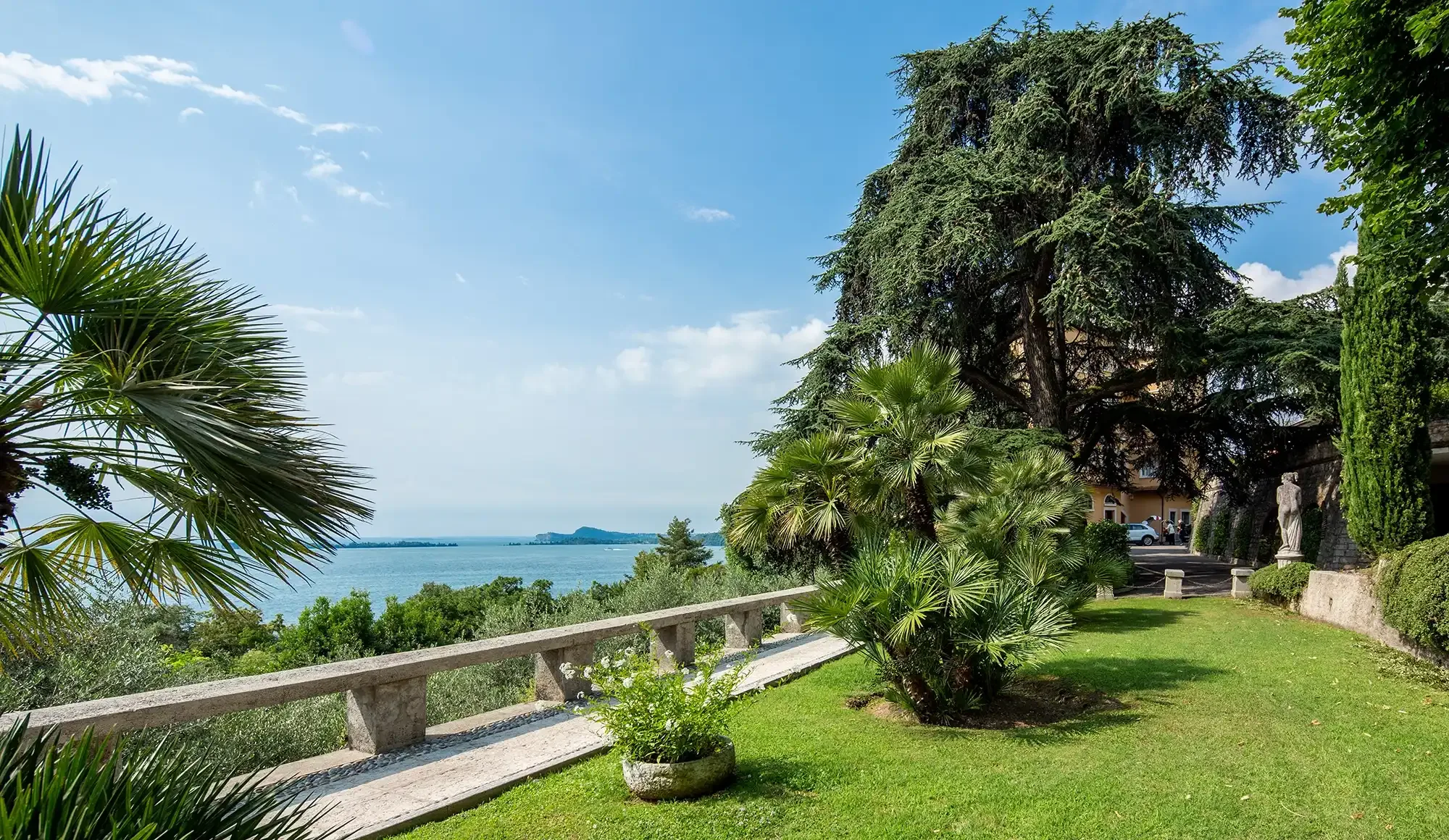 Giardino con palme e balaustra affacciato su lago azzurro e colline